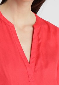 Donna che indossa una blusa rossa con scollatura a V e un dettaglio cucito nella parte inferiore della scollatura.
