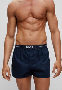 Navy blue swim shorts with elastic waistband, featuring a Boss logo and a button closure. Made from smooth, lightweight material.