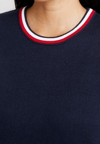 Pull en maille bleu marine avec un col rond. Le col côtelé présente des rayures rouges et blanches. Texture lisse avec un poids moyen.