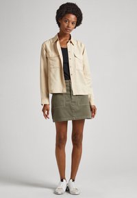 Cămașă bej cu butoni, cu două buzunare la piept, purtată peste un top negru, asortată cu o fustă verde și teniși albi. Design simplu, look casual.