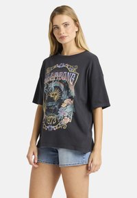 Ξανθιά γυναίκα που φοράει ένα oversize μαύρο t-shirt Billabong με ήλιο, φοίνικες, λουλούδια και εκτύπωση του 1973, σε συνδυασμό με ανοιχτόχρωμο τζιν σορτς.