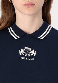 Tamsiai mėlynas polo marškinėliai su baltu juostuotu apykakle ir ryškiu baltu Hilfiger logotipu, pasižymintys tekstūruotu audiniu.