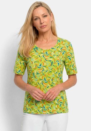 Femme blonde portant une chemise vert citron à manches courtes avec un motif tourbillonnant coloré et un pantalon blanc, debout les mains jointes lâchement.