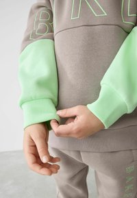 Grauer Pullover mit hellgrünen Ärmeln und Bündchen. Auf der Vorderseite ist das Wort "BAKE" in Grün aufgedruckt, mit weicher Textur und sportlicher Passform.