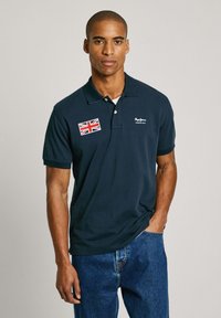 Marineblauw poloshirt met kraag, voorzien van een geborduurd Union Jack-patch en logo aan de linkerkant. Gemaakt van gladde stof.