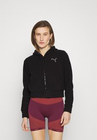 Felpa nera corta con zipper, con logo Puma bianco. Abbinata a pantaloni corti color bordeaux, messi in risalto da un motivo a righe e una vita abbinata.