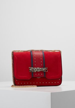 Sac à main en daim rouge avec bandoulière chaîne dorée, bordure noire, détails cloutés et fermeture en forme de tête de léopard dorée sur une surface blanche.