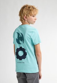 Camiseta de algodón de color azul claro con cuello redondo, que presenta gráficos de llamas y engranajes de color azul oscuro en la parte posterior, y el texto "PETROL IND" en la parte inferior.