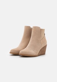Bottes de cheville en daim beige avec un talon compensé, dotées d'un bout pointu et de panneaux latéraux côtelés pour un enfilage facile.