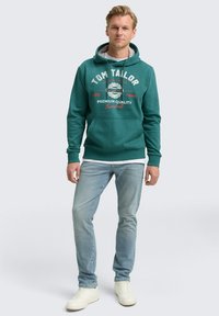 Sudadera color teal con un bolsillo frontal, capucha con cordón y gráfico impreso. Combinada con jeans azul claro y zapatillas blancas. Ajuste casual.