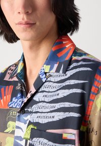 Short-sleeve shirt in vibrant colors featuring a collage of patterns, including foliage and text, with a relaxed collar and pocket detail.