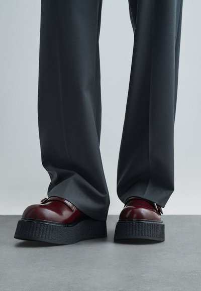 Σκούρο γκρι παντελόνι με φαρδιές γάμπες πάνω σε γυαλιστερά μπορντό πλατφόρμες, τοποθετημένο σε γκρι πάτωμα μπροστά από ομοιόμορφο φόντο.