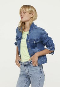 Jeansjacke in mittlerem Blau mit Stehkragen, zwei Brusttaschen und silbernen Knöpfen. Getragen über einem hellgrünen T-Shirt.