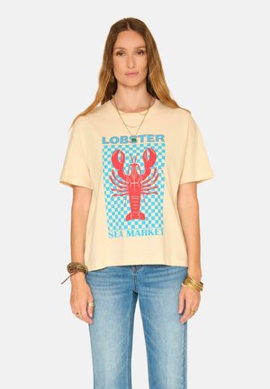 Femme portant un t-shirt beige avec un graphique rouge de homard et un motif à carreaux bleu, associé à un jean bleu clair et des bijoux en or, debout devant un fond blanc.