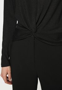 Svart långärmad topp med en vriden frontdetalj, tillverkad av ett glittrande material, tillsammans med enkla svarta byxor. Slät yta och figursydd design.
