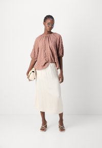 Braune, strukturierte Bluse mit gerafftem Ausschnitt, kombiniert mit einem schwingenden cremefarbenen Rock. Die Accessoires bestehen aus einer hellen Handtasche und beigen Sandalen mit Riemchen.