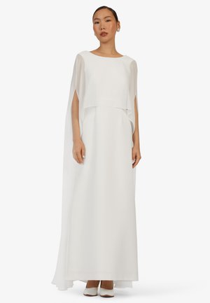 Frau trägt ein bodenlanges weißes Kleid mit fließenden Kapuzenärmel und weißen Absätzen, steht vor einem schlichten hellen Hintergrund.