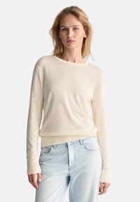 Maglione beige chiaro realizzato in un tessuto morbido, con scollo rotondo e maniche lunghe con polsini a costine, abbinato a jeans blu chiaro.