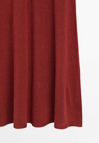 Partie d'une jupe en tissu couleur rouille avec un tricot doux et texturé suspendu devant un fond blanc.