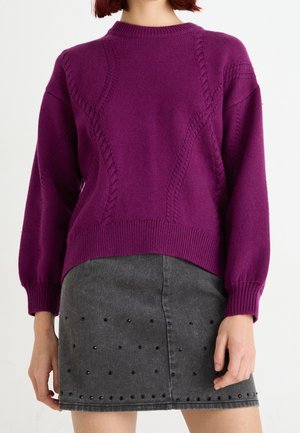 Femme portant un pull violet à manches longues en maille torsadée et une jupe gris foncé avec des ornements cloutés noirs.