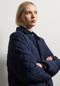 Donna bionda con le braccia incrociate, indossando una giacca trapuntata blu navy, che guarda pensierosa di lato su uno sfondo chiaro e semplice.