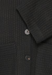 Tissu noir à côtes avec des coutures verticales, comprenant deux boutons noirs et un détail de poignet replié.