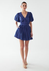 Ženska v temno modri obleki z napihnjenimi rokavi, V-izrezom, nabranim pasom in valovitim robom, obuta v bež sandale s odprtimi prsti.