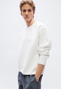 Λευκό φούτερ με στρογγυλή λαιμόκοψη και ριμπ μανσέτες, με διακριτικό ανάγλυφο λογότυπο στο μπροστινό μέρος. Συνδυάζεται με γκρι υφασμάτινο παντελόνι.