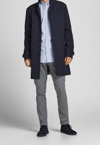 Marinblå kappa med skräddarsydd passform, knappstängning fram och sidofickor, bärs över en ljusblå skjorta med knappar och grå strukturerade byxor.