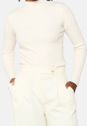 Persona que lleva un suéter de cuello alto de canalé color crema de manga larga metido dentro de pantalones de cintura alta color crema, con las manos en los bolsillos.
