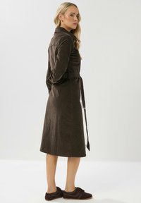 Braunes, knielanges Kleid aus einem samtartigen Material, mit tailliertem Gürtel, langen Ärmeln und einem Hemdkragen. Kombiniert mit dunklen Turnschuhen.