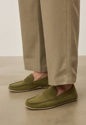Pieds portant des mocassins en daim vert olive associés à un pantalon beige raccourci sur un sol taupe lisse.