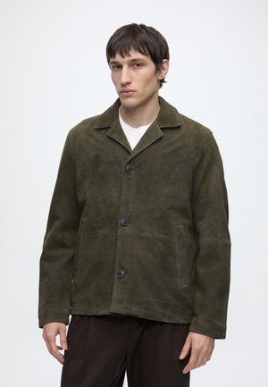 Homme portant une veste en daim vert foncé sur une chemise blanche à boutons noirs, debout devant un fond clair uni.