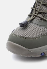 Πεζοπορικό παπούτσι σε ελαιί πράσινο με πλέγμα και συνθετικά υλικά, στρογγυλεμένη μύτη, ελαστικά κορδόνια και λαστιχένια σόλα με μικρή μπλε λεπτομέρεια.