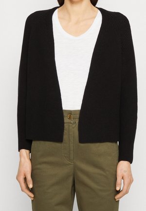 Femme portant un cardigan en tricot noir ouvert sur un haut blanc à col en V et un pantalon taille haute vert olive avec fermeture à boutons à l'avant.
