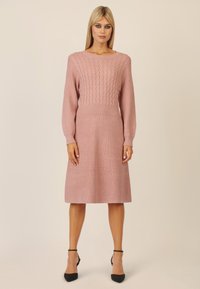 Gestricktes Kleid in sanftem Pink mit gerippter Textur und Zopfmuster im Oberteil. Mit langen Ärmeln und einem ausgestellt geschnittenen Rock, kombiniert mit schwarzen Heels.