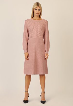 Gestricktes Kleid in sanftem Pink mit gerippter Textur und Zopfmuster im Oberteil. Mit langen Ärmeln und einem ausgestellt geschnittenen Rock, kombiniert mit schwarzen Heels.