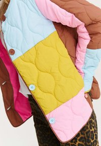 Prešívaná mnohofarebná bunda s hnedými, ružovými, žltými a modrými časťami a gombíkmi, nosená cez oranžové nohavice s leopardím vzorom.