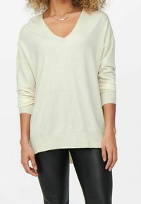 Femme portant un pull crème à col en V avec des manches longues et un pantalon en cuir noir, accessoirisée avec un collier et des bagues.