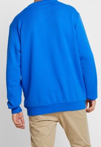 Sudadera azul de corte holgado, mangas largas, puños y dobladillo acanalados. Textura suave y diseño minimalista. Combinada con pantalones beige.