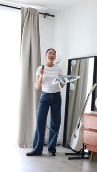 Ung kvinde i hvid t-shirt og jeans holder pakket tøj og en sort genstand, stående ved et gulv-til-loft spejl og en hvid guitar.
