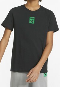 Maglietta nera a maniche corte con patch del logo Puma verde che presenta un design di volto pixelato, indossata da una persona con pantaloni grigi chiari a vestibilità ampia.