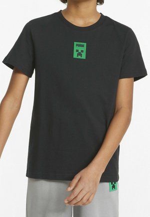 Musta lyhythihainen paita, jossa on vihreä Puma-logopatch, jossa on pikselöity kasvosuunnittelu, päällä henkilö, jolla on päällään löysät vaaleanharmaat housut.