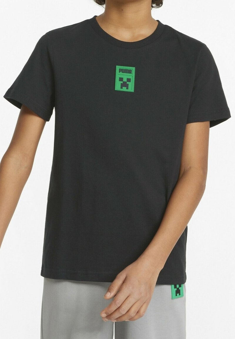 Maglietta nera a maniche corte con patch del logo Puma verde che presenta un design di volto pixelato, indossata da una persona con pantaloni grigi chiari a vestibilità ampia.