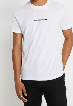 T-shirt en coton blanc avec un col rond, arborant un logo noir "CALVIN KLEIN" sur la poitrine, assorti à un pantalon noir.