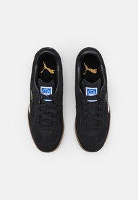 Schwarze Wildleder-Sneaker mit einer Sohle aus Gummi, ausgestattet mit Schnürsenkeln, weißen Seitenstreifen und goldenen Logo-Akzenten. Flaches, abgerundetes Zehendesign.