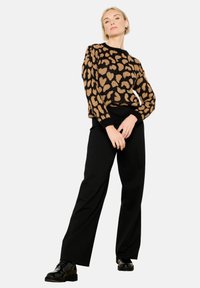 Strickpullover mit schwarzem Grund und beigem Leopardenmuster, kombiniert mit schwarzen weit geschnittenen Hosen und schwarzen Schuhen.