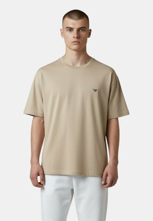 Giovane uomo con capelli corti che indossa una maglietta beige con un piccolo logo sul petto e pantaloni bianchi, in piedi contro uno sfondo semplice.