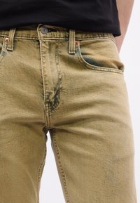 Κοντινό πλάνο σε μπεζ τζιν από τζιν ύφασμα που φοριούνται με μια μαύρη πουκαμίσα που είναι βιδωμένη μέσα, δείχνοντας τις μπροστινές τσέπες, το κουμπί και τις θηλιές της ζώνης.