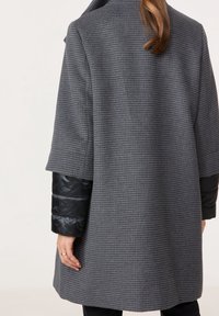 Šedý vlněný kabát s texturovaným vzorem houndstooth. Má černé nadýchané rukávy a volný střih. Bez viditelných zapínání.